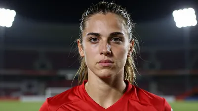 A focused mid-20s female footballer in a vibrant red jersey, tight head-and-shoulders crop