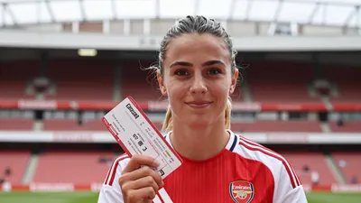 A young, anonymous Arsenal Women player in a red jersey with white sleeves, holding a ticket and smiling, with a blurred...