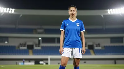 A 25-year-old female footballer in a blue jersey with white sleeves and a white collar, wearing a pair of white shorts...