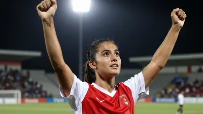 A young, athletic female footballer in a red and white kit, celebrating a goal with arms raised