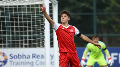 A 19-year-old German winger with short, dark hair and a determined expression, wearing a bright red jersey with white...