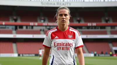 A tight, waist-up shot of an anonymous Arsenal Women player in a white jersey with a red collar, wearing a determined...